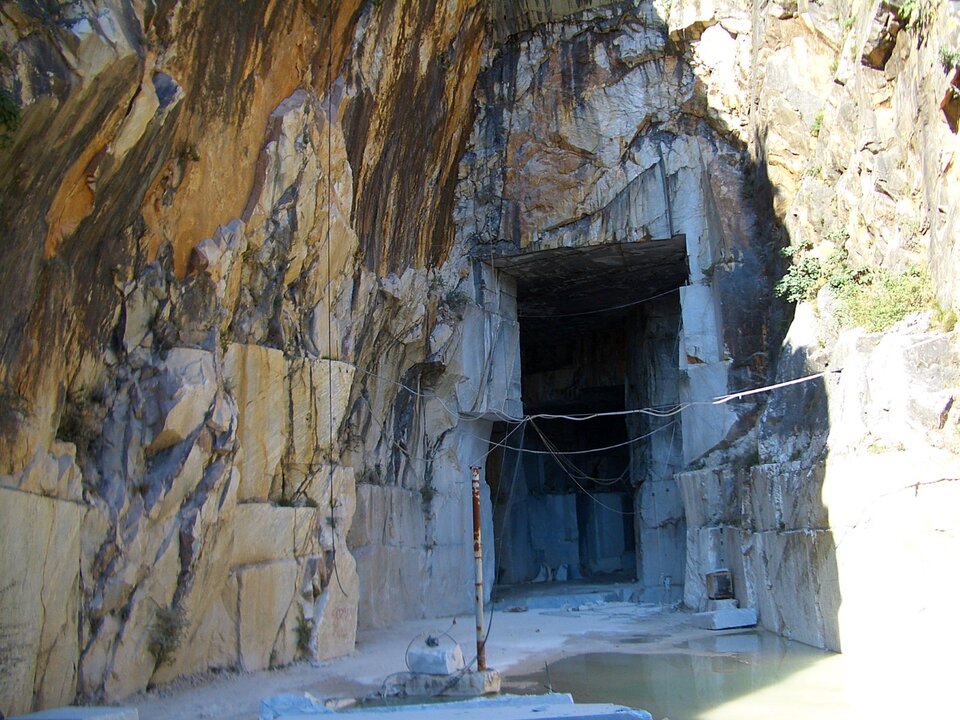 Ilustracja przedstawia skały marmurowe w Carrarze. W części środkowej zdjęcia widoczny jest otwór, który powstał przez wycięcie bloków skalnych. Fotografia została wykonana w słoneczny dzień - część skał jest w cieniu, część w intensywnym słońcu.