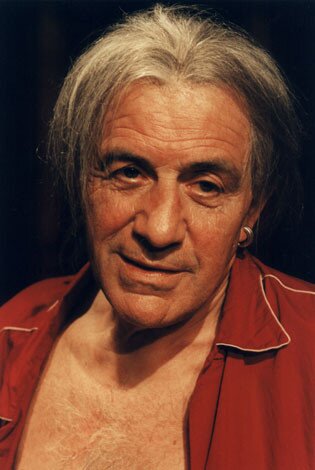 Na zdjęciu widać twarz mężczyzny, który ma lekko rozczochrane dłuższe siwe włosy. W lewym uchu ma srebrny kolczyk w kształcie koła. Ubrany jest w czerwoną rozpiętą piżamę. Ma rozmarzony wyraz twarzy.