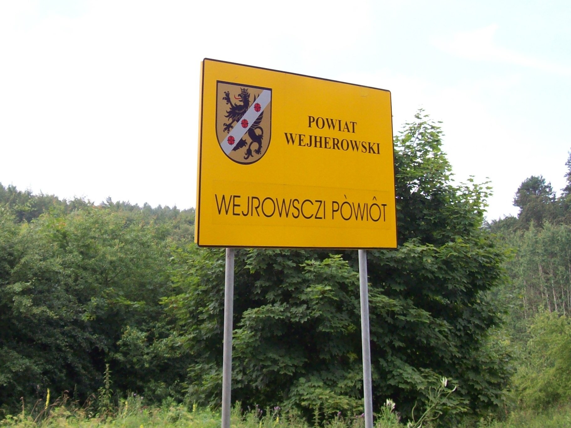 Kliknij, aby powiększyć Zdjęcie. Tablica informacyjna stoi przy drodze. Na żółtym tle znajduje się herb Wejherowa. Tarcza herbowa ma złote pole, gdzie znajduje się czarny gryf przepasany srebrną wstęgą, na której widnieją trzy czerwone róże. Obok herbu widnieje czarny drukowany napis powiat wejherowski. Poniżej została zapisana w języku kaszubskim informacja wejrowsczi powiot. Tablica stoi w pobliżu lasu.