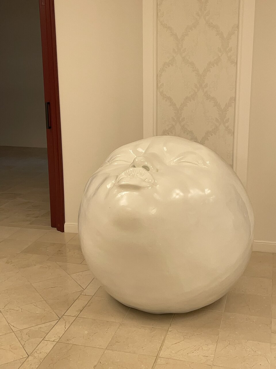 Ilustracja przedstawia rzeźbę umieszczoną na podłodze we wnętrzu. Rzeźba ma kształt kuli, na której wyraźnie zaznaczona jest twarz skierowana ku górze z zamkniętymi oczami. Powierzchnia rzeźby jest gładka i połyskująca.