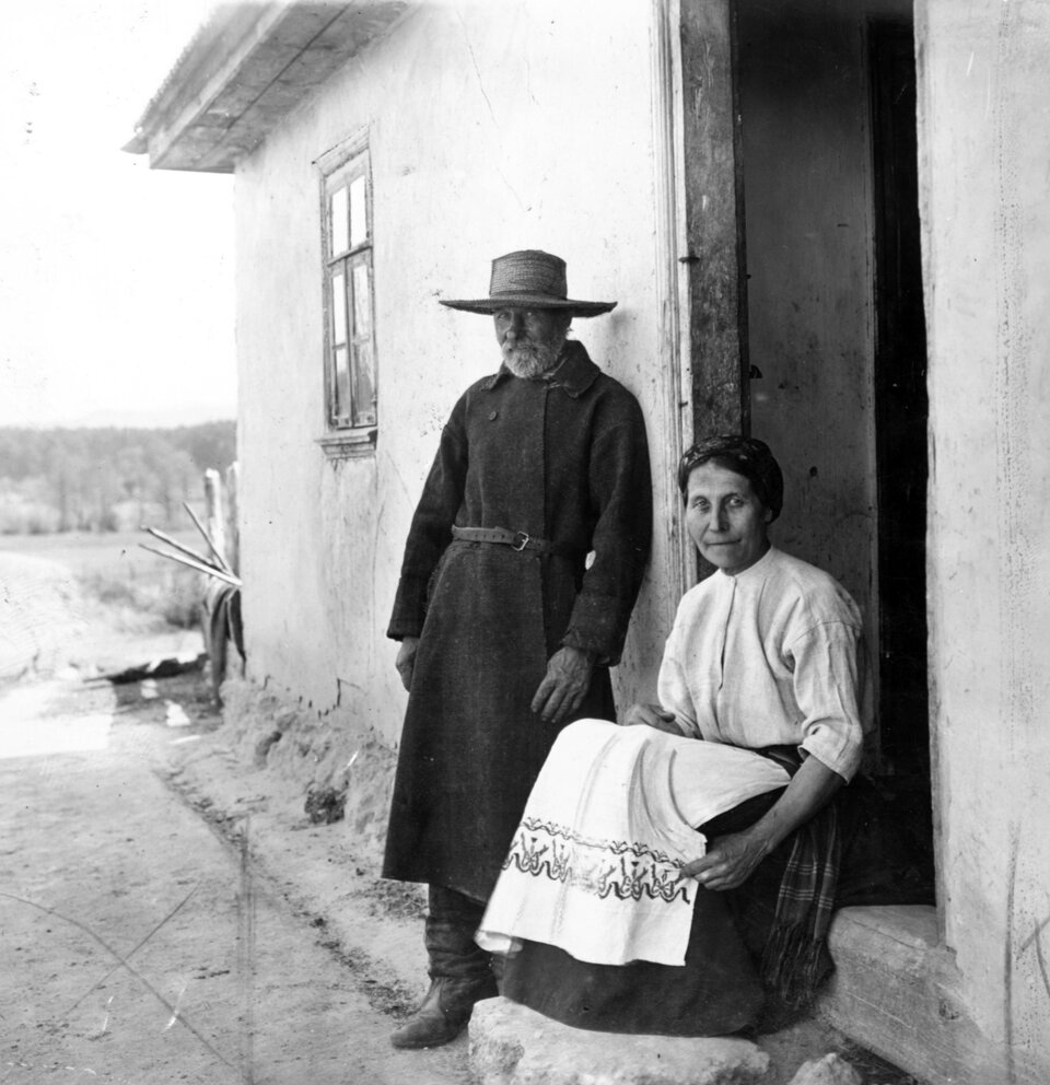 Zdjęcie przedstawia mieszańców Wołynia. Kobieta siedzi na progu chaty. Jest ubrana w białą bluzkę oraz w spódnicę z tradycyjnym wzorem na dole. Obok oparty o ścianę stoi mężczyzna. Ma na sobie długi ciemny płaszcz do ziemi. W pasie przewiązany jest paskiem. Na głowie ma kapelusz. Chata ma jasne ściany. W tle są pola.
