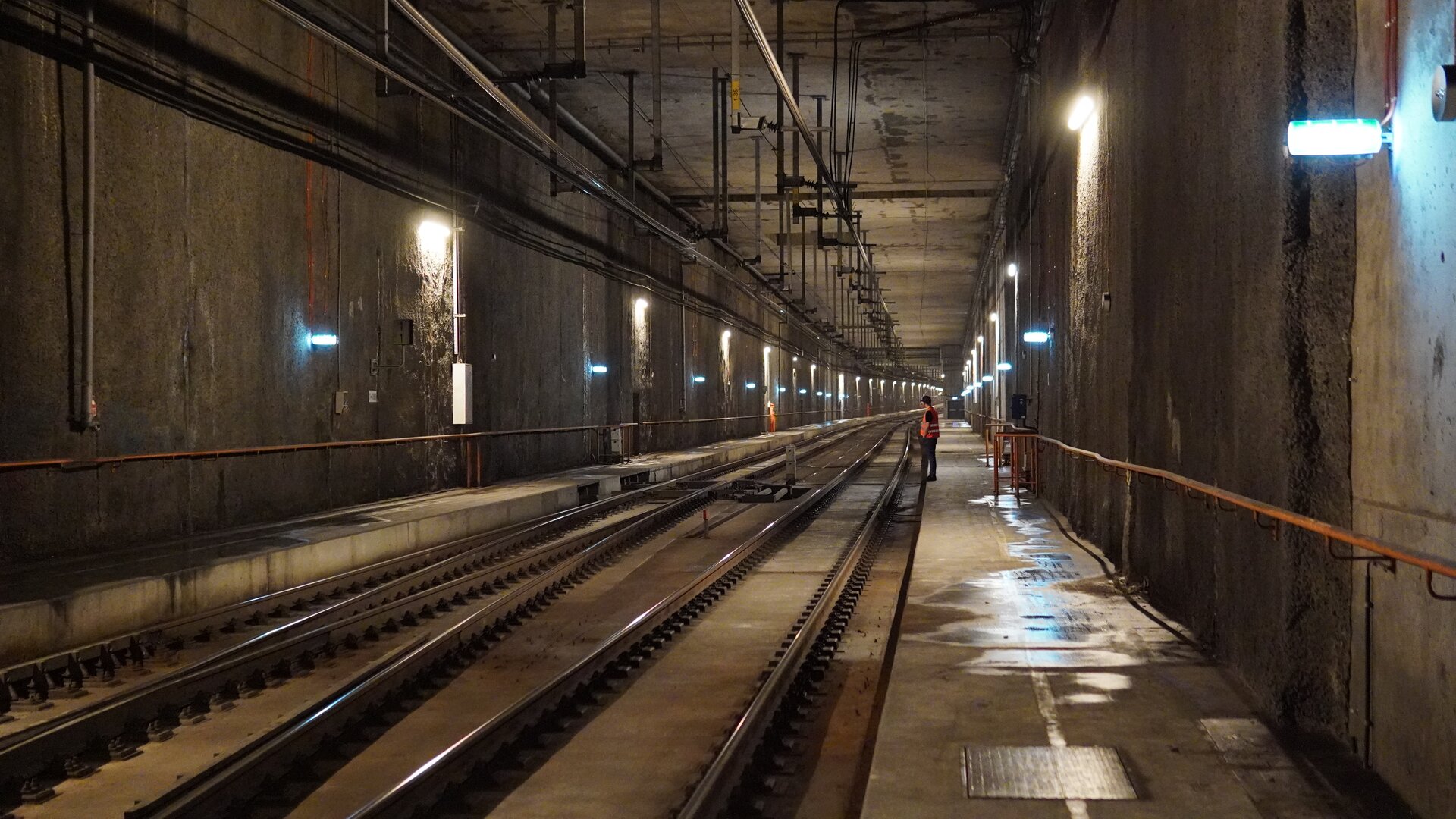 Ilustracja przedstawia tunel kolejowy wykonany z betonu. Wnętrze tunelu jest dobrze oświetlone, urządzenia oświetlające zamontowane są na jego ścianach. W tle widoczne jest dwutorowe torowisko oraz pracownicy kolei w odblaskowych kamizelkach, którzy prawdopodobnie wykonują prace na linii kolejowej. Obok torowiska znajdują się chodniki, z których korzystają pracownicy. Na suficie tunelu widoczne są wiszące metalowe elementy.