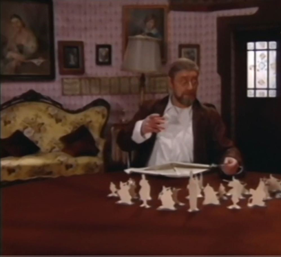 Zdjęcie przedstawia dojrzałego mężczyznę z okularami, ciemnosiwymi włosami i brodą z wąsami, który siedzi przy drewnianym stole. Ubrany jest w białą koszulę i brązową marynarkę. Na stole znajdują się papierowe figurki.