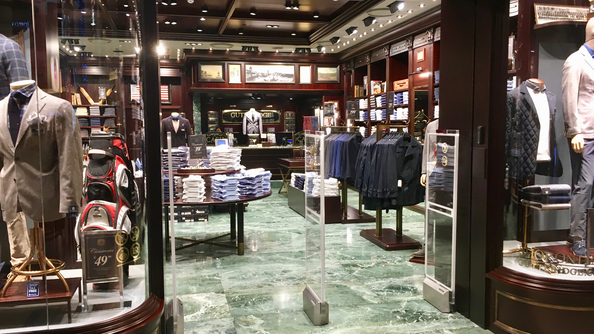 Zdjęcie przedstawia wejście oraz wnętrze męskiego, eleganckiego sklepu odzieżowego. Na witrynie stoją manekiny ubrane w garnitury, marynarki i eleganckie kurtki. Wewnątrz sklepu, na stole widoczne są ułożone koszule. Na wieszakach wiszą marynarki oraz płaszcze. Na półkach leżą koszule. 