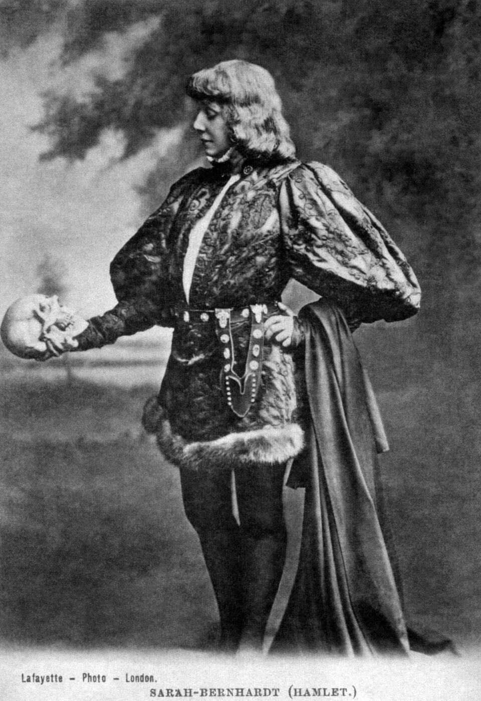 Zdjęcie przedstawia aktorkę, Sarah'e Bernhardt, w roli Hamleta. Kobieta ma na sobie szesnastowieczny strój męski, W prawej dłoni trzyma ludzką czaszkę.