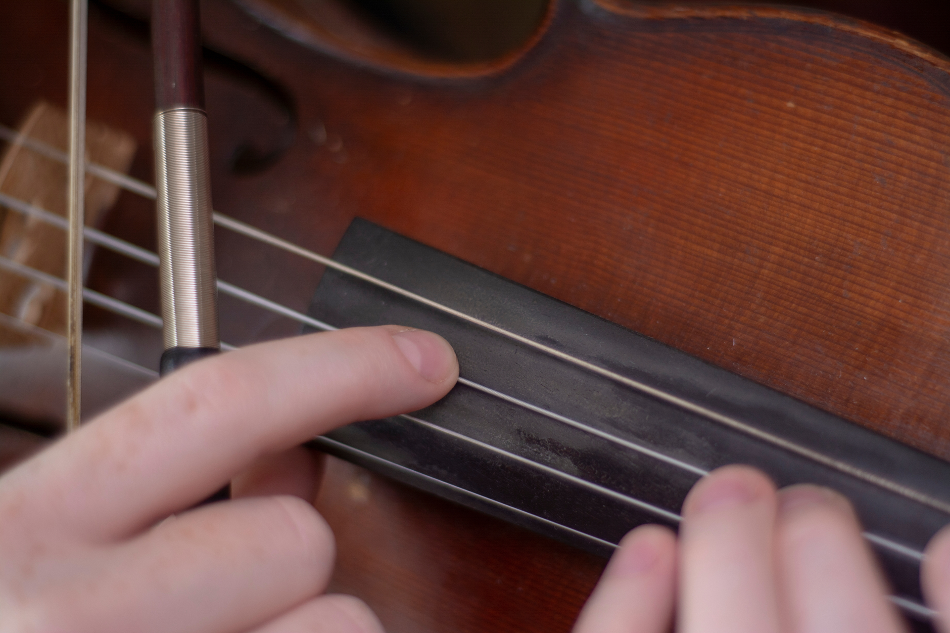 Fotografia przedstawia technikę pizzicato [czytaj: piccikato] na skrzypcach. Pokazane jest zbliżenie na fragment drewnianych skrzypiec oraz trzymające je dwie dłonie na czarnym gryfie z czterema srebrnymi strunami. Instrument ten jest w ciemnobrązowym kolorze. Po lewej stronie zdjęcia w jednej dłoni, osoba trzyma srebrno‑brązowy smyczek, a wskazującym palcem przyciska od góry, drugą strunę.