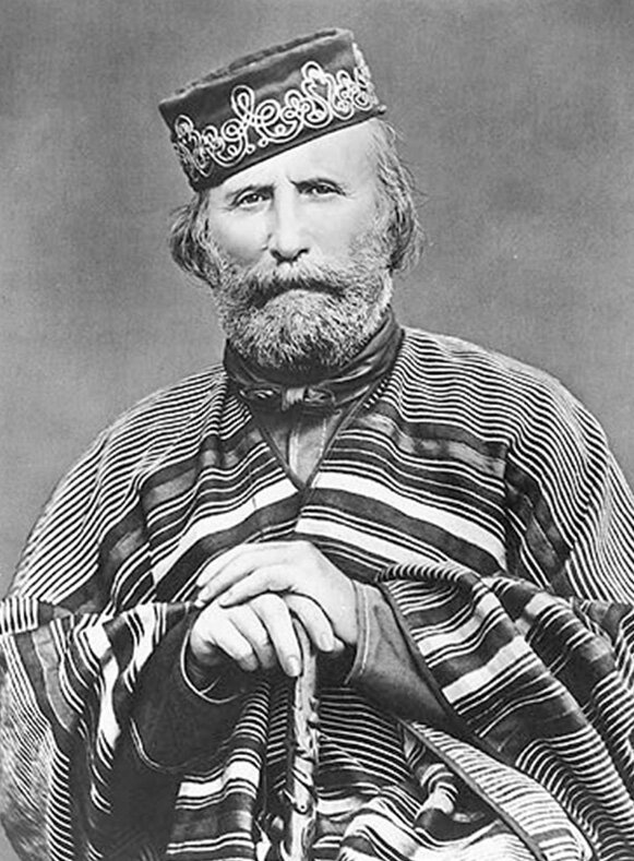 Fotografia czarno-biała przedstawiająca Giuseppe Garibaldiego. Brodaty mężczyzna w narzucie w pasy. Ma elegancko wyszyte nakrycie głowy, dość duży zarost na twarzy. Obydwie dłonie trzyma oparte na lasce. Pozuje do zdjęcia.