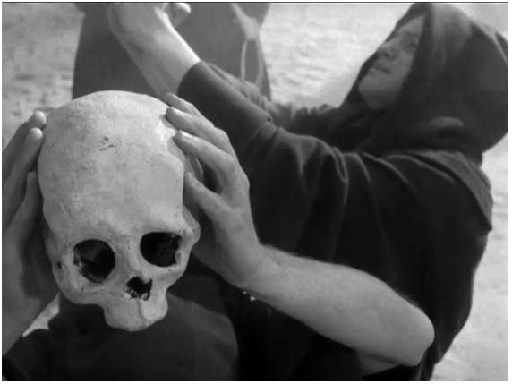 Fotografia przedstawia męskie dłonie trzymające ludzką czaszkę. Na drugim planie ukazany jest zakapturzony mężczyzna. Unosi ręce.