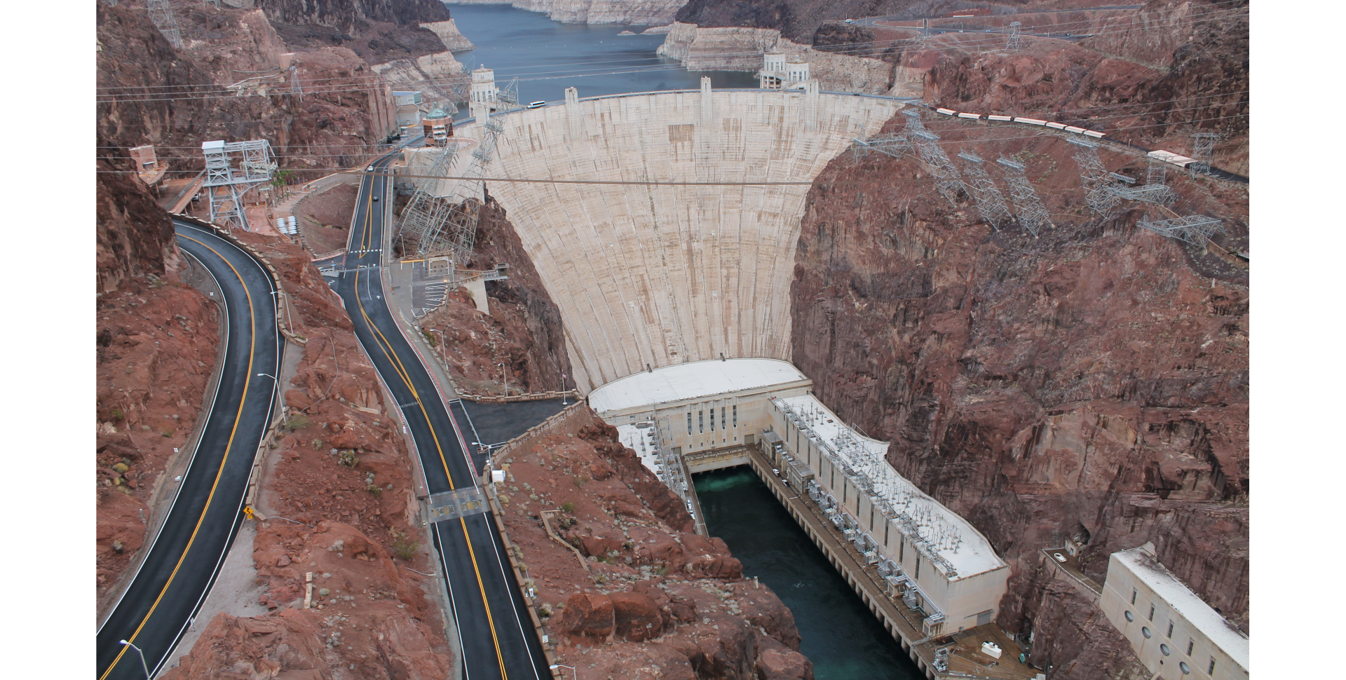 Zdjęcie. Widok hydroelektrowni. Pośrodku tama wykonana z betonu zatrzymująca umieszczoną wyżej wodę zgromadzoną w zbiorniku. Poniżej tamy budynki hydroelektrowni, sieć energetyczna. Z lewej strony drogi asfaltowe prowadzące do hydroelektrowni.