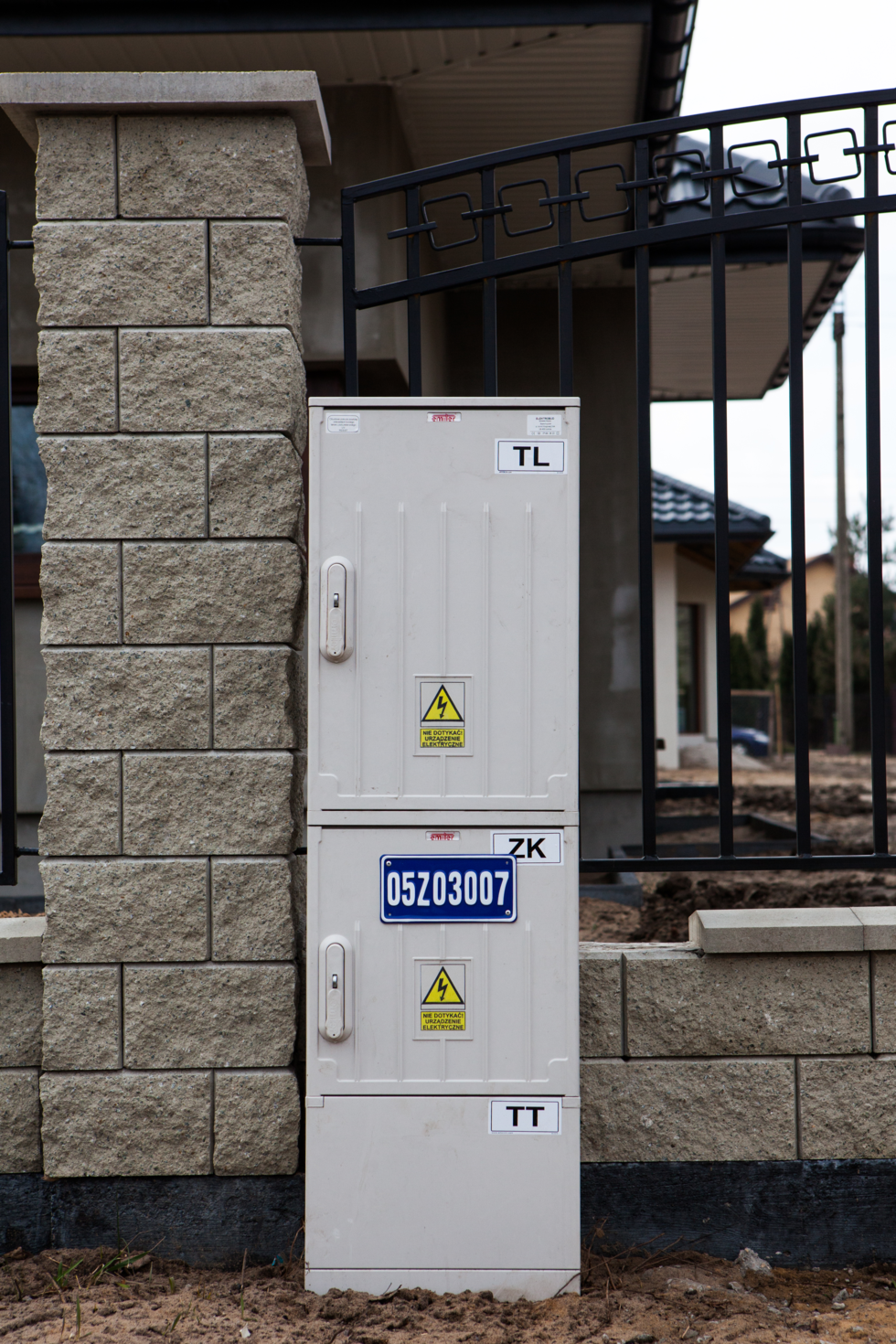 Fotografia przedstawia metalową popielatą skrzynię metalową przyczepioną do metalowego płotu przydomowego. W tle widać fragmenty domów, osiedla domków jednorodzinnych.