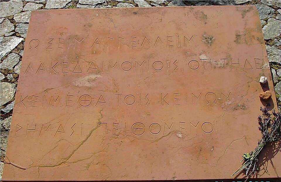 Fotografia przedstawia kamienną płytę pamiątkową z napisem wyrytym greckimi literami. Płyta ma kolor miedziany; znajduje się na podłożu składającym się z szarych kamieni. 