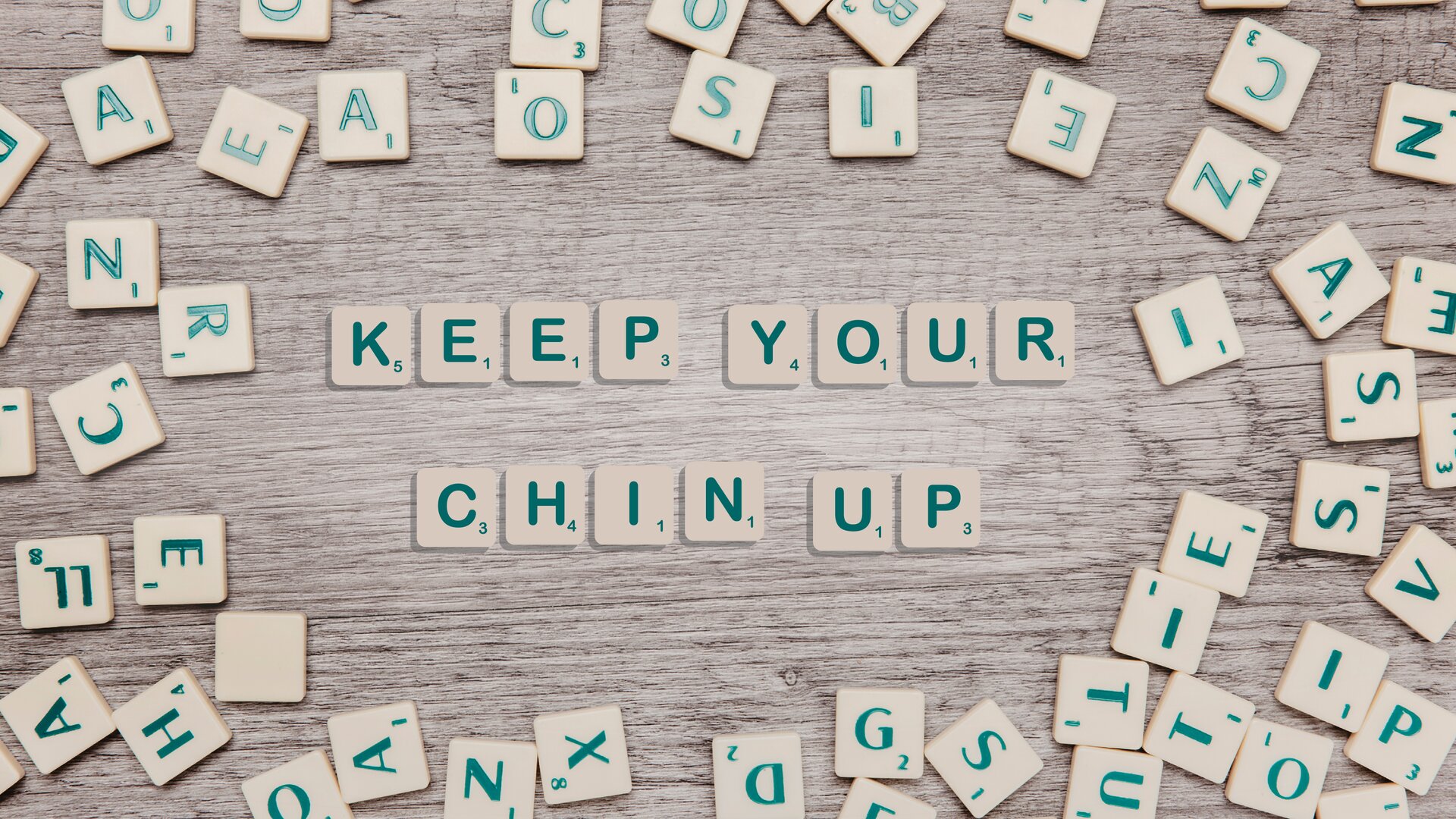 Zdjęcie przedstawia rozrzucone na drewnianym blacie kremowe kwadraciki z zielonymi literkami oraz małymi numerami po prawej, dolnej stronie każdej litery. Pośrodku ułożone zostały cztery słowa w języku angielskim: KEEP YOUR CHIN UP.