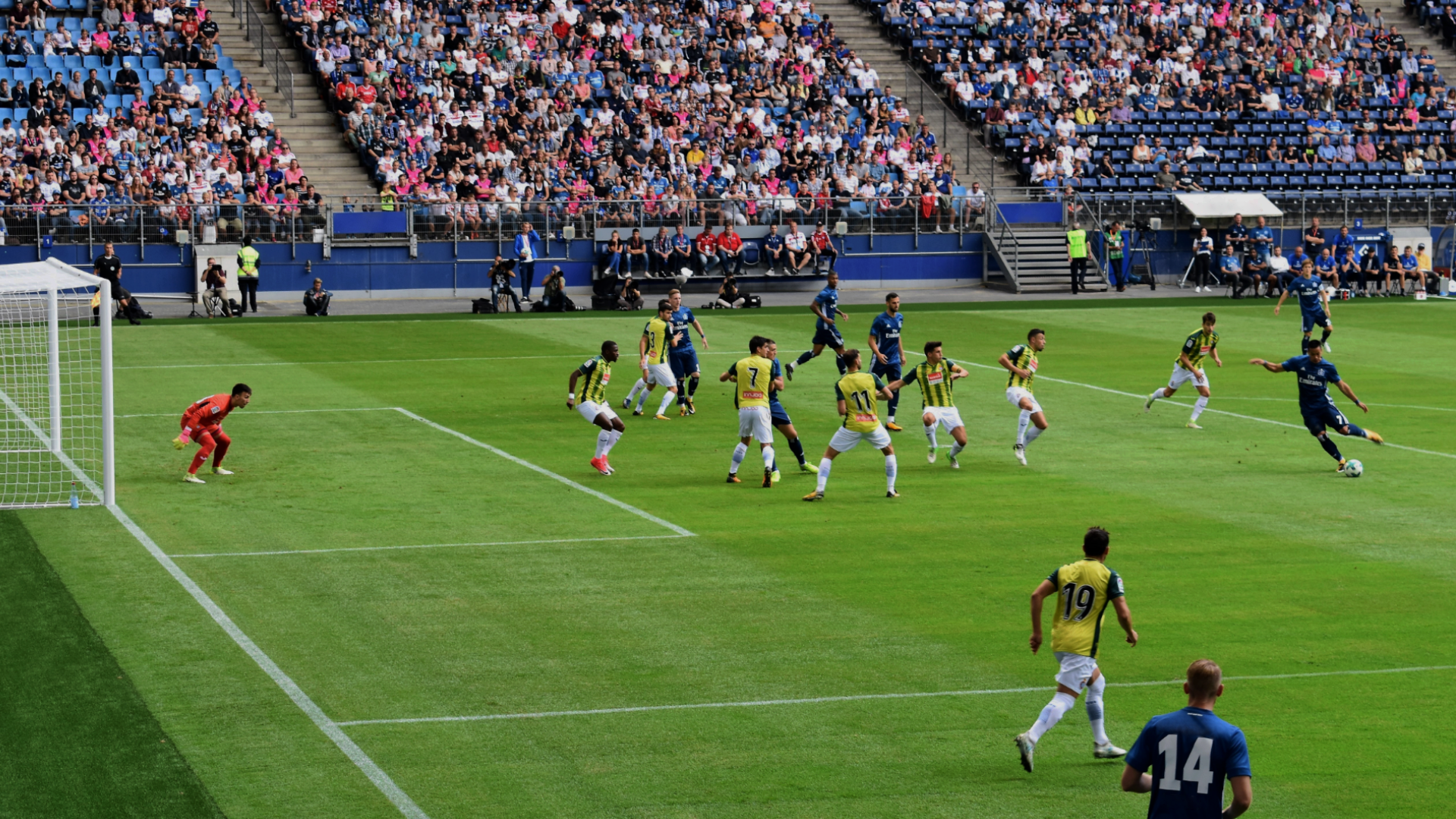 Zdjęcie przedstawia mecz piłki nożnej rozgrywany na stadionie. Widoczni są piłkarze na murawie i kibice na trybunach.