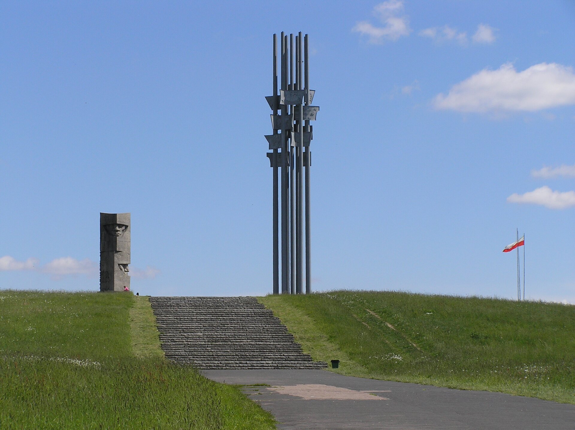 Na zdjęciu widoczne są łąka i schody prowadzące do pomników grunwaldzkich. Pierwszy pomnik stanowią wysokie metalowe pręty, drugi zaś jest kamienny i widać jego fragment. Po prawej stronie w oddali na maszcie powiewa polska flaga. W tle widoczne jest niebieskie niebo. 