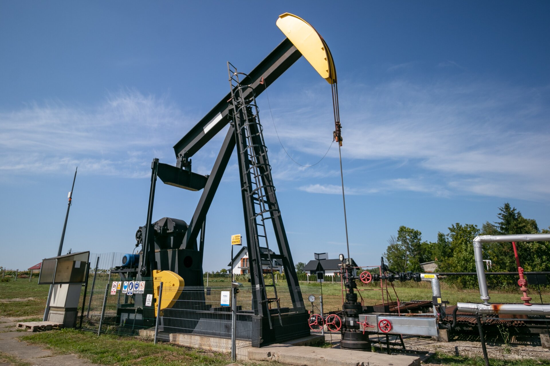 Na zdjęciu widać czarno‑żółty indywidualny żuraw pompowy wraz z infrastrukturą napowierzchniową otworu. Żuraw jest to urządzenie wprawiające w ruch przewód pompowy a poprzez przewód pompę wgłębną.Widoczny w centralnej części zdjęcia żuraw jest wykonany z czarnej ramy o kształcie ostrosłupa o podstawie trójkąta. Na szczycie ramy osadzone jest ramie żurawia zakończone z prawej strony głowicą. Ramie wraz z żółtą głowicą jest w kształcie młotka. Do głowicy przymocowana jest cienki czarny drążek polerowany który łączy się z czarną głowicą eksploatacyjną. Po drugiej stronie ramie znajduje się przeciwwaga o kształcie trapezu do którego boków przymocowane są dwie belki o kształcie podłużnych prostokątów czyli pociągacze. Pociągacze połączone są z czarnym silnikiem. Po prawej stronie głowicy widoczne są dwie szare rury.W tle zdjęcia widać dwa trzy obiekty mieszkalne które charakteryzują się spadzistym dachem.