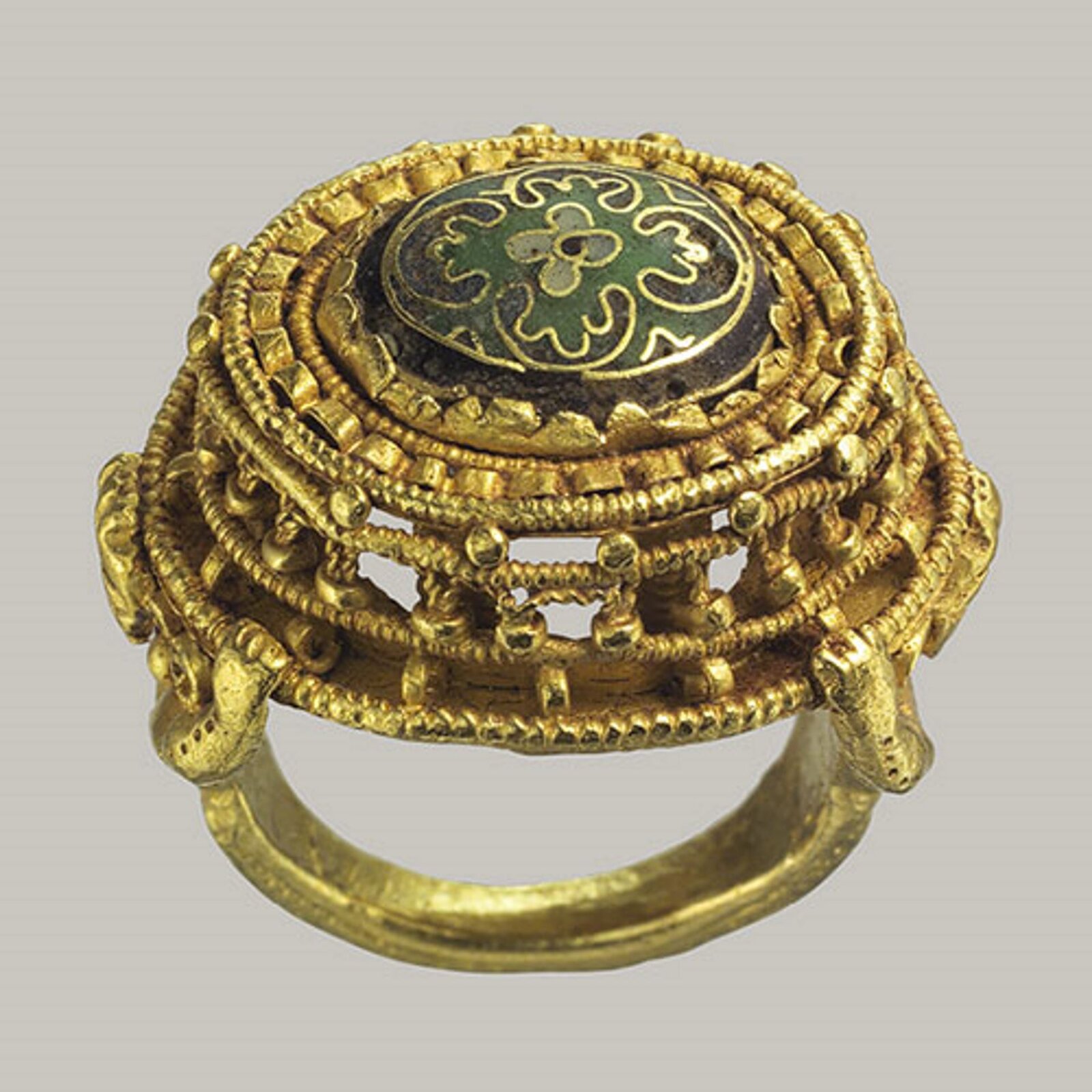 Fotografia przedstawia pozłacany pierścień, który składa się z wielu elementów ze sobą zlutowanych. Tworzą go liczne okręgi, zmniejszające się ku górze i wstęgi giętego drutu. Pośrodku znajduje się kwiatowy ornament z emalii Cloisonné, jest to zielony kwiatek o złotych brzegach i nieregularnych płatkach.