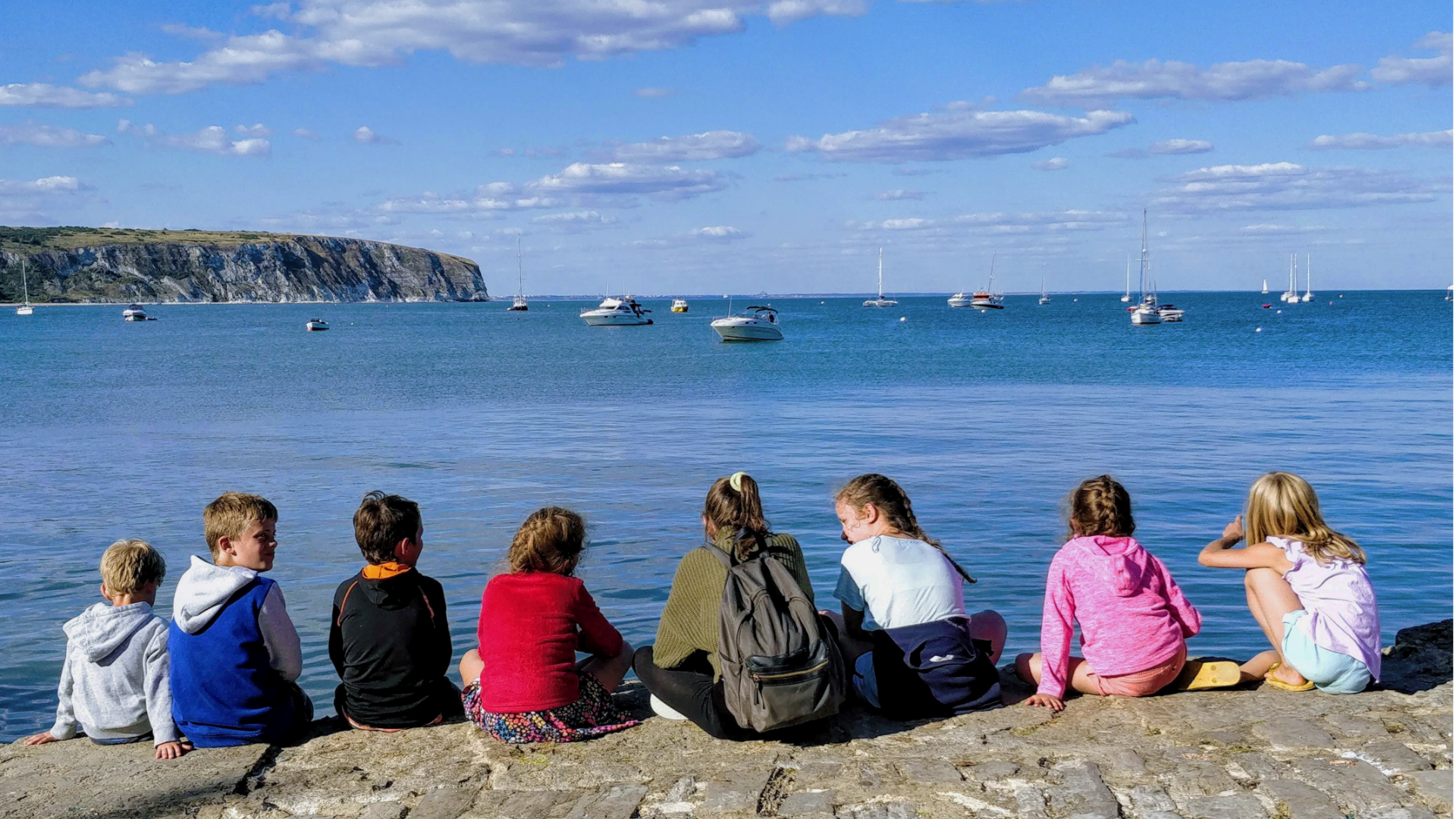 Zdjęcie przedstawia grupę dzieci siedzących nad brzegiem morza. Dzieci siedzą tyłem do kadru.