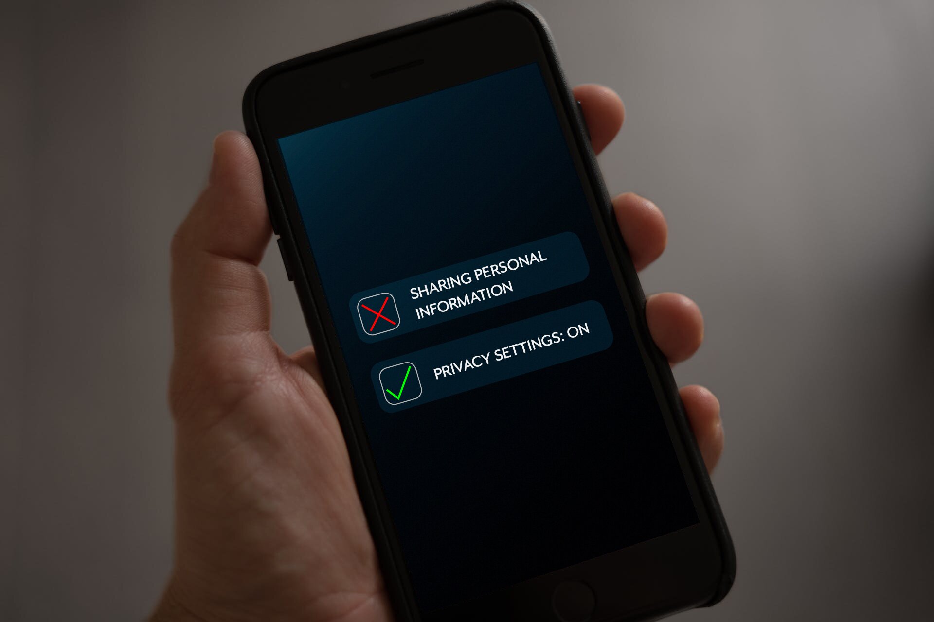 Zdjęcie przedstawia telefon komórkowy typu smartfon, trzymany w lewej dłoni. Na czarnym ekranie telefonu widoczne są dwa napisy: górny – SHARING PERSONAL INFORMATION, po lewej stronie którego widoczny jest kwadracik z czerwonym iksem w środku; oraz dolny – PRIVACY SETTINGS: ON, po lewej stronie którego widoczny jest kwadracik z zielonym znakiem ptaszka w środku.