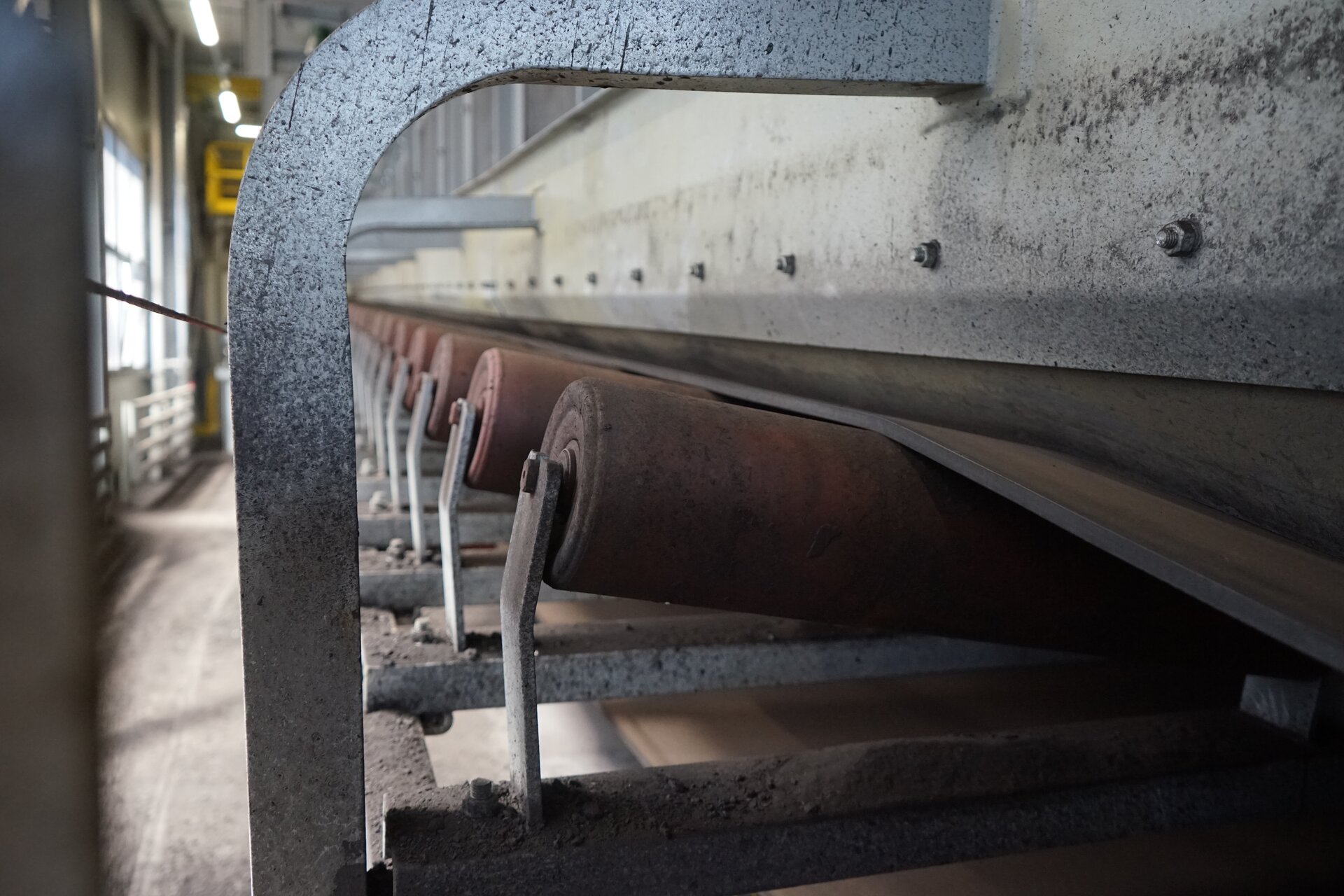 Na zdjęciu widać fragment przenośnika taśmowego z jednym z jego podstawowych podzespołów czyli krążnikiem. Krążniki są to obrotowe wałki, po których przesuwa się taśma przenośnika.Przenośnik taśmowy jest to podłużne urządzenie mechaniczne na górze którego znajduje się czarna taśma. Podstawą przenośnika taśmowego jest stalowa konstrukcja kratowa czyli trasa. Konstrukcja ta zbudowana jest z powtarzalnych członów modułów których podstawą są ceowniki. Ceownik jest to wyrób hutniczy, walcowany lub gięty z metalu. Wyroby te są w postaci prostych odcinków. Przekrój poprzeczny czyli profil ceownika przypomina literę C.Z boku konstrukcji czyli na boku trasy znajdują się metalowe elementy w kształcie ceowników, które biegną pionowo w górę. Końcówki tych elementów są zakrzywione w kierunku taśmy czyli w kierunku środka przenośnika. Są to kozły górne. Kozły górne są rozmieszczone po oby stronach trasy. Na koźle górnym widać krążnik górny. Krążnik jest w kształcie czerwonego walca. Na krążnikach znajduje się długa taśma która biegnie przez całą długość przenośnika.Od boku trasy odchodzą równomiernie stalowe elementy  w kształcie odwróconej litery L. Przedstawiony na zdjęciu przenośnik znajduje się we wnętrzu stalowej hali.