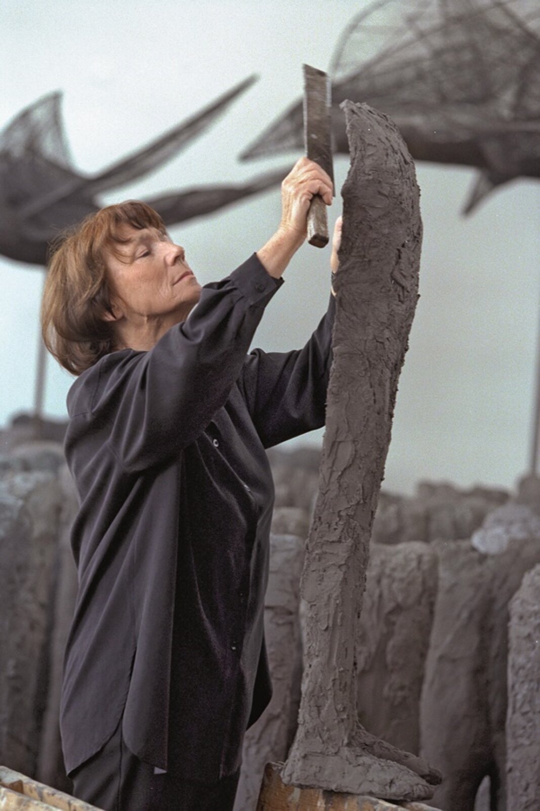 Fotografia przedstawia Magdalenę Abakanowicz podczas prac nad rzeźbą. Kobieta w średnim wieku ma średniej długości brązowe włosy z grzywką opadającą na czoło. Na jej twarzy widoczny jest delikatny uśmiech. Ubrana jest w luźną ciemną koszulową bluzkę oraz czarne spodnie. Kobieta stoi z lewej strony a rzeźba z prawej i przedstawia ona długie brązowe nogi o chropowatej fakturze. W jednej dłoni trzyma narzędzie z trzonem oraz prostokątnym zakończeniem, a w drugiej fragment rzeźby, którą docina. W tle widoczne są pozostałe fragmenty rzeźby – nogi, na jasnym tle.
