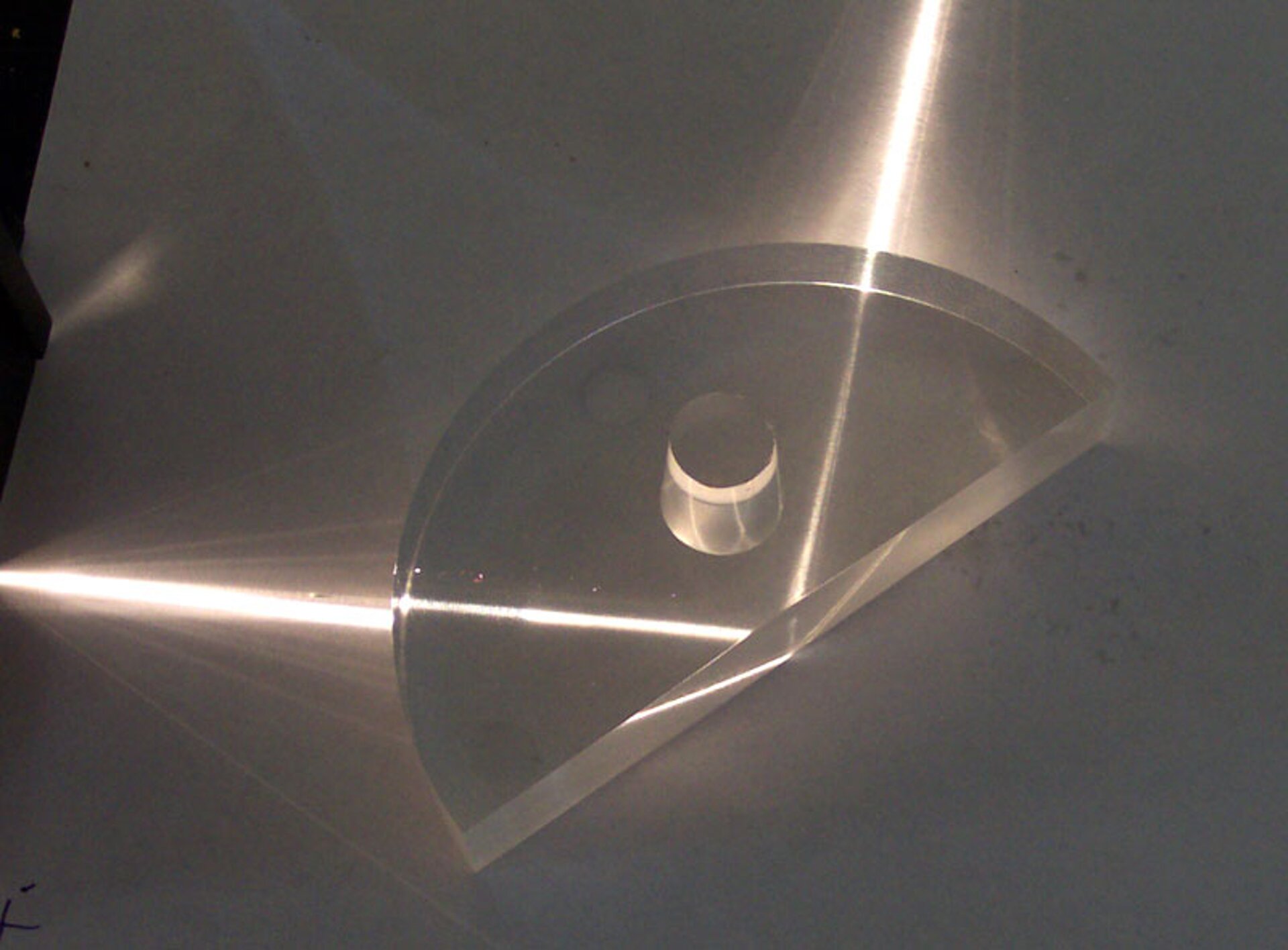 Rys. 3. Zdjęcie przedstawia od góry szklany element w kształcie połowy płaskiego walca podzielonego pionową płaszczyzną przechodzącą przez oś symetrii walca. Na powierzchnię boczną walca pada promień światła laserowego skierowany wzdłuż promienia walca. Promień przechodzi bez załamania przez kolistą ścianę boczną walca i pada pod małym kątem na płaską powierzchnię połówki walca. Promień odbija się całkowicie od granicy szkła z powietrzem. Promień odbity ma prawie taką samą jasność jak promień padający. 