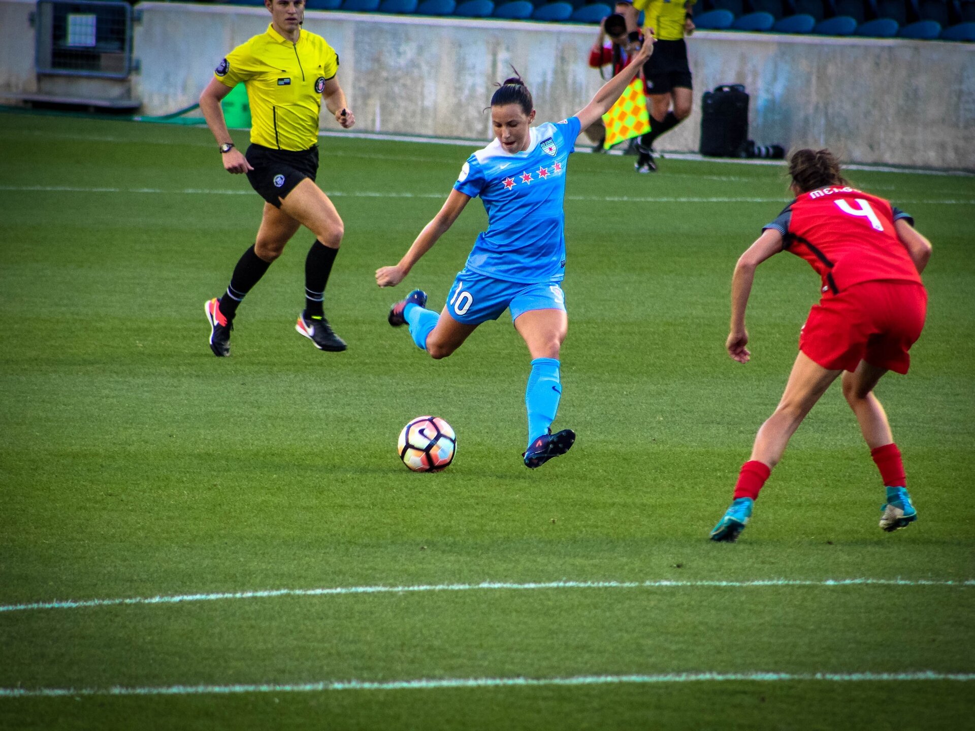 Zdjęcie przedstawia mecz kobiet w piłkę nożną. Widać dwie zawodniczki zróżnych zespołów w tym jedna w natarciu oraz sędziego. W oddali znajduje się sędzia boczny oraz reporter.