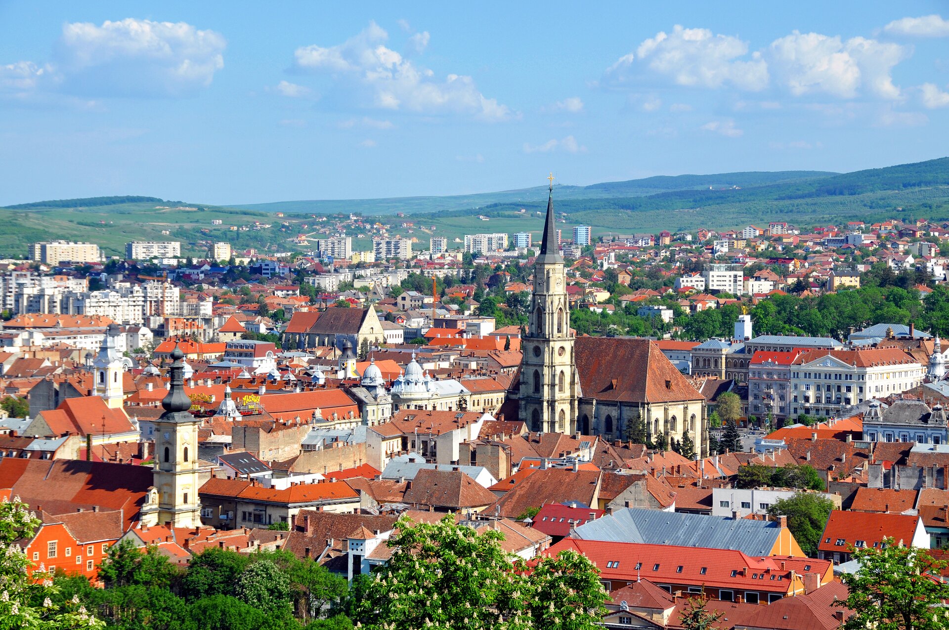 Zdjęcie przedstawiające zabudowania miejsce, kamienice w kolorze czerwonym, pośrodku widoczna wieża. W tle pagórki i wzniesienia.