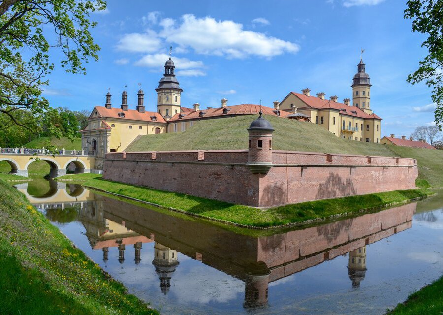  Fotografia  przedstawiająca turystyczną atrakcję Białorusi - Zamek w Nieświeżu. Zamek zbudowany jest z cegły , otoczony murem obronnym oraz fosą. Do zamku prowadzi most. 