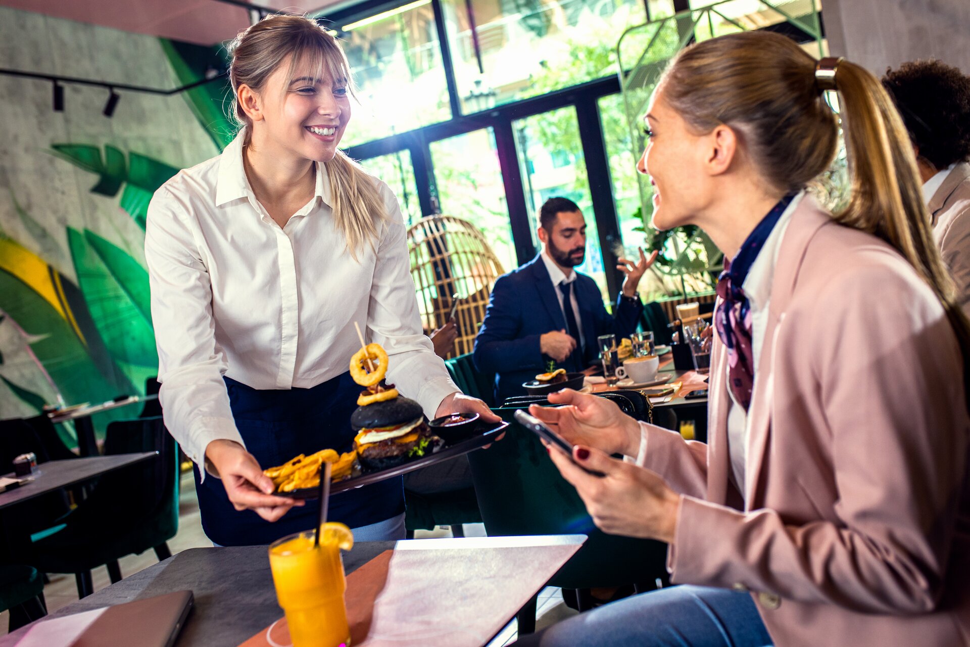 Na zdjęciu widać scenę z restauracji, gdzie uśmiechnięta kelnerka w białej koszuli i czarnym fartuchu podaje danie do stolika. Klientka w różowym swetrze, siedząca przy stoliku, wydaje się być w dobrych humorach, patrzy na telefon i coś mówi. Na tacy, którą trzyma kelnerka, znajduje się burger z czarną bułką oraz frytki.