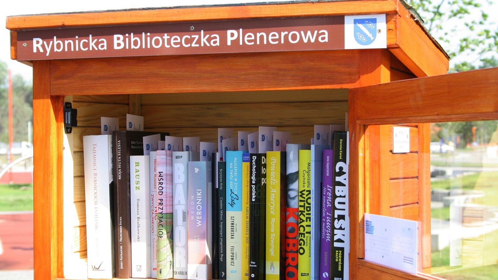 Zdjęcie ukazuje drewnianą, brązową skrzynkę z książkami stojącą w plenerze. 