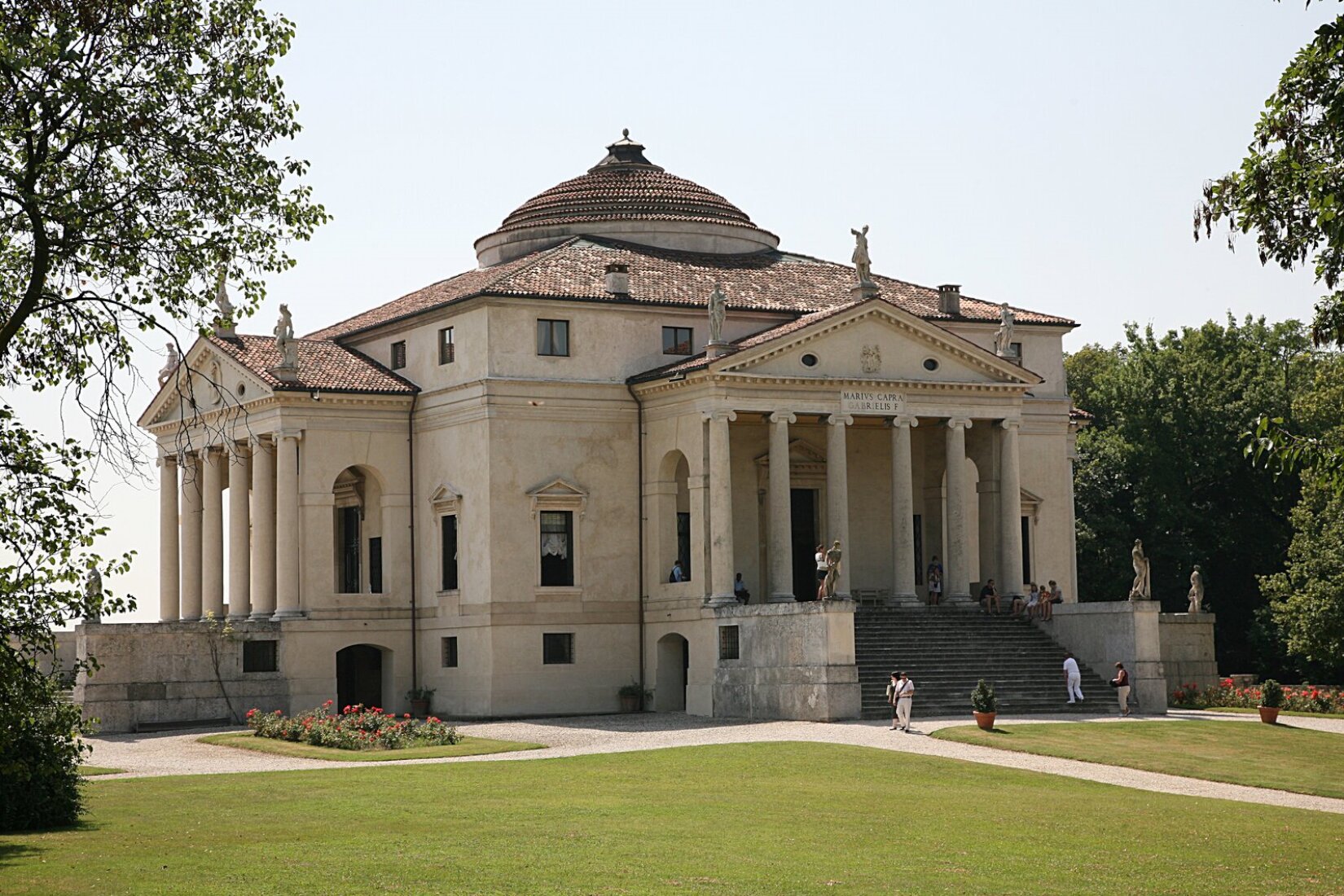 Fotografia przedstawia budynek, do którego prowadzą schody. Przed głównym wejściem znajdują się kolumny wraz z portykiem. Na fryzie budynku znajduje się napis, który brzmi: MARIUS CAPRA GABRIELIS F. Powyżej znajduje się kopuła. Na lewym oraz prawym skrzydle budynku, jak i na kopule znajdują się posągi. Przed budynkiem rosną dwa krzewy oraz zielona trawa.