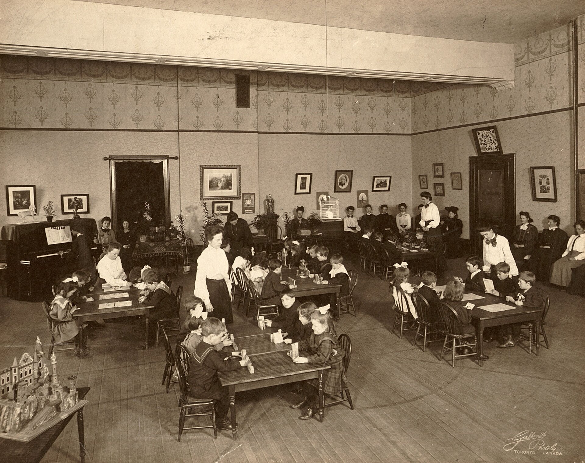  Na zdjęciu dzieci siedzące przy stolikach w przedszkolu. Stolików z dziećmi jest pięć, na szóstym stoi makieta budynku. Każdym ze stolików opiekuje się jedna nauczycielka. Zależnie od stolika dzieci czytają, układają klocki, jedzą. Wokół, przy ścianach, siedzą dorosłe kobiety, obserwujące ich. Jedna z kobiet gra na pianinie. Na ścianach zawieszone obrazy.   