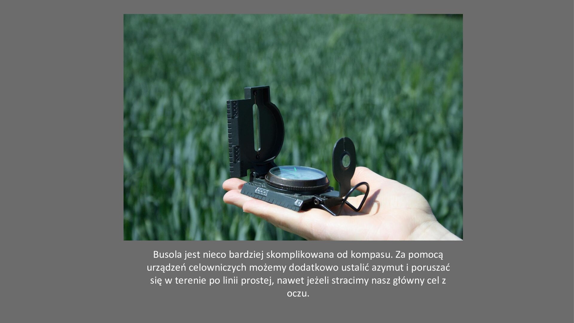 Szary slajd. Na środku zdjęcie przedstawiające dłoń ludzką, na której leży czarna busola. W tle zielona łąka. Pod zdjęciem tekst: „Busola jest nieco bardziej skomplikowana od kompasu. Za pomocą urządzeń celowniczych możemy dodatkowo ustalić azymut i poruszać się w terenie po linii prostej, nawet jeżeli stracimy nasz główny cel z oczu”