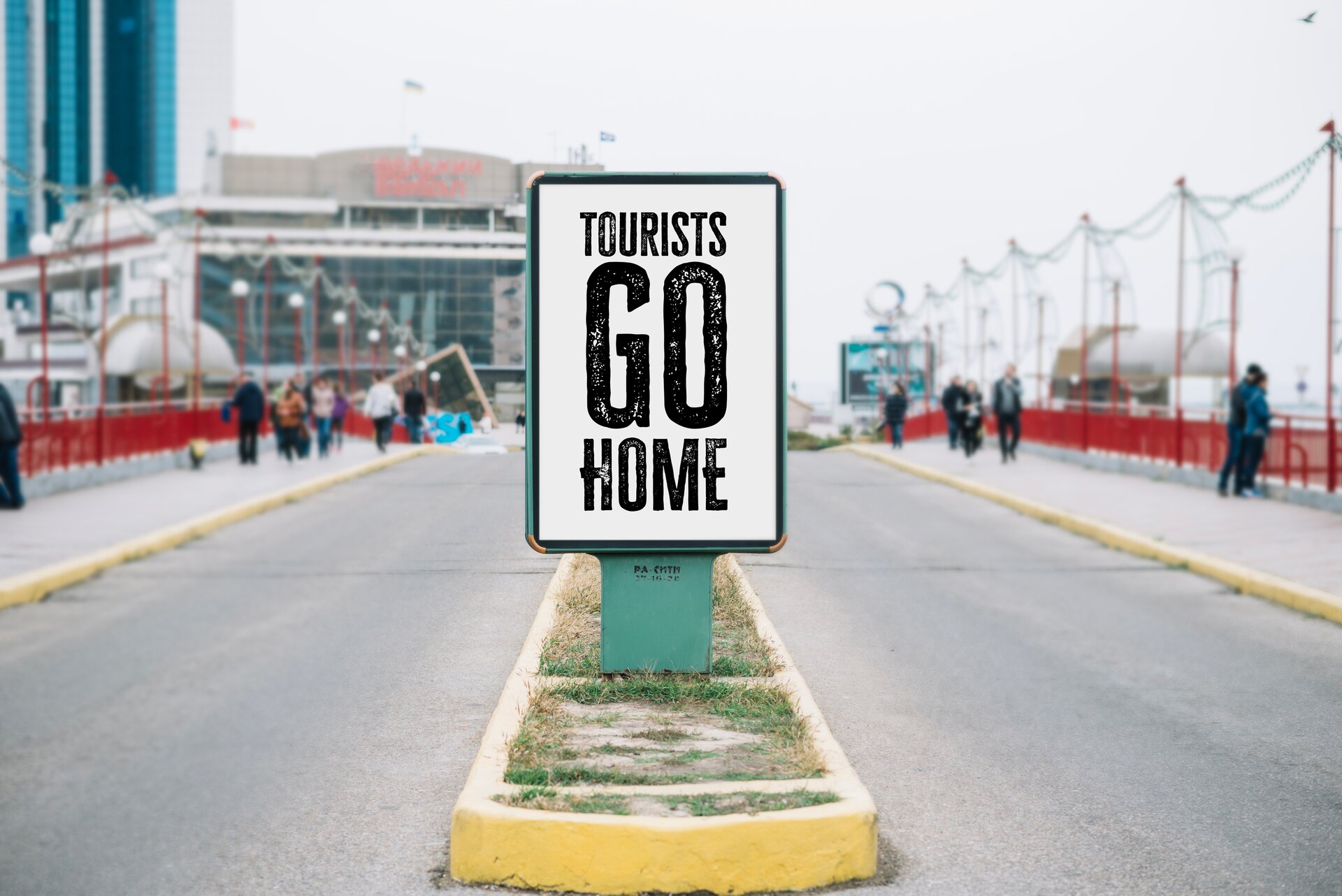 Zdjęcie przedstawia ulicę, która jest przedzielona wzdłuż małym pasem trawnika, wokół którego jest krawężnik. Na trawniku dzielącym pasy jezdni jest tablica z napisem "TOURISTS GO HOME". Transkrypcja do nagrań znajduje się poniżej.