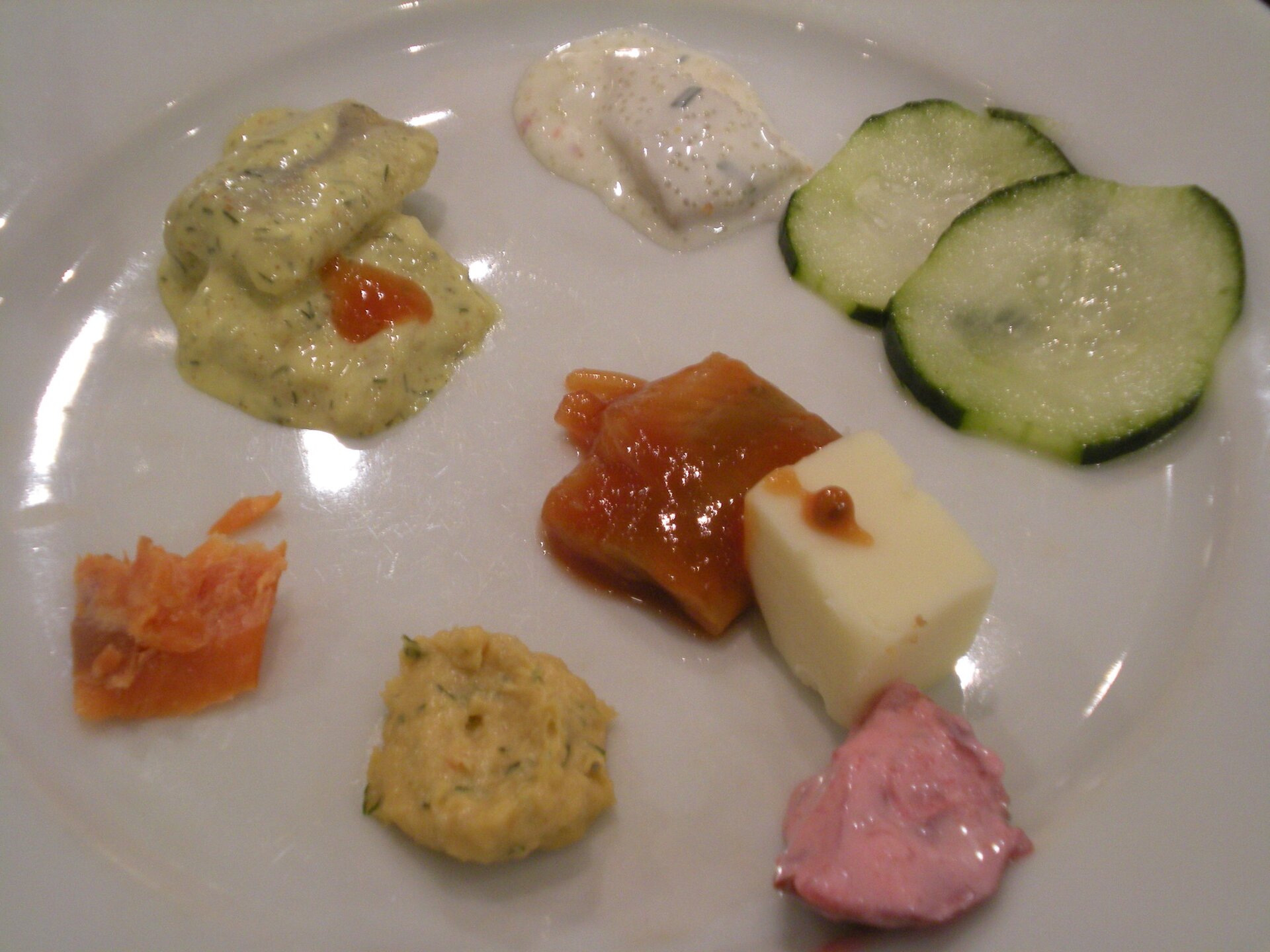 Kliknij aby powiększyć Na zdjęciu widoczny biały talerz (widok z góry), na którym leżą sześć małych kęsów jedzenia, mały kawałek masła i dwa plasterki ogórka.