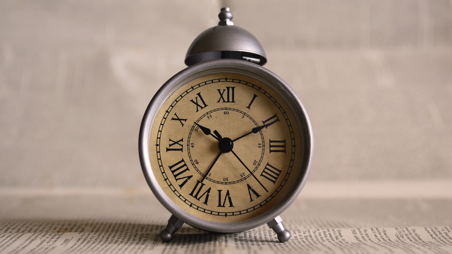 Zdjęcie przedstawia okrągły budzik retro. Na górze budzika znajduje się grzybek z dzwonkiem oraz przycisk do wyłączania dzwonka. Godziny na tarczy zegara oznaczone są rzymskimi cyframi. Zegar wskazuje godzinę 10:11. Budzik ustawiony jest na gazecie.