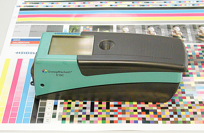 Zdjęcie przedstawia densytometr. Jest to średniej wielkości urządzenie o lekko prostopadłościennym kształcie. Na wierzchniej jego części znajduje się wyświetlacz, umożliwiający użytkownikowi odczyt wyników. Urządzenie znajduje się na zadrukowanej próbkami koloru płachcie papieru.