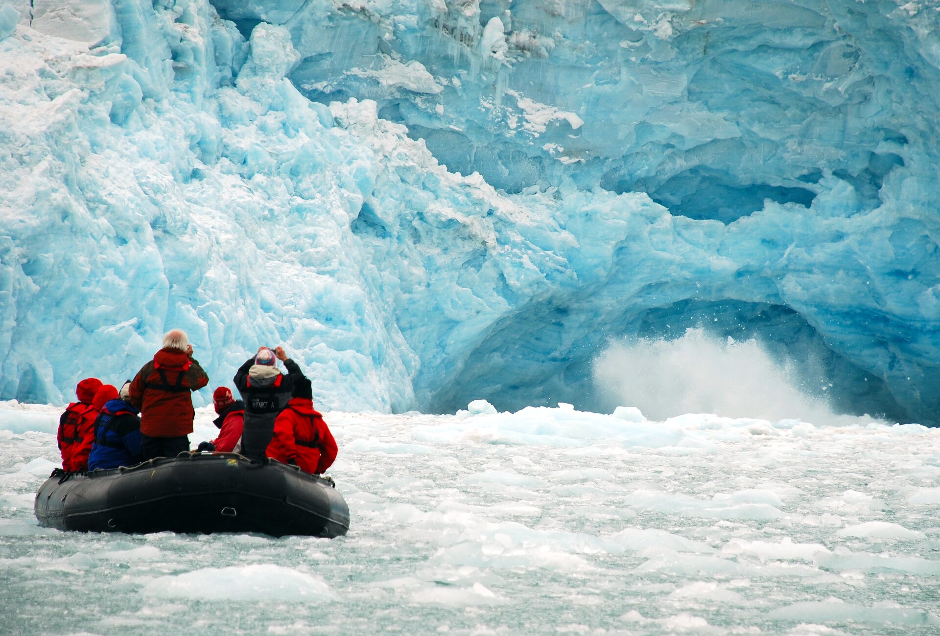 Na zdjęciu lodowiec schodzący bezpośrednio do wód morskich. Widać kawałku lodu w ruchu. Spiętrzona woda pokryta krą, ludzie w pontonie fotografują czoło lodowca.
