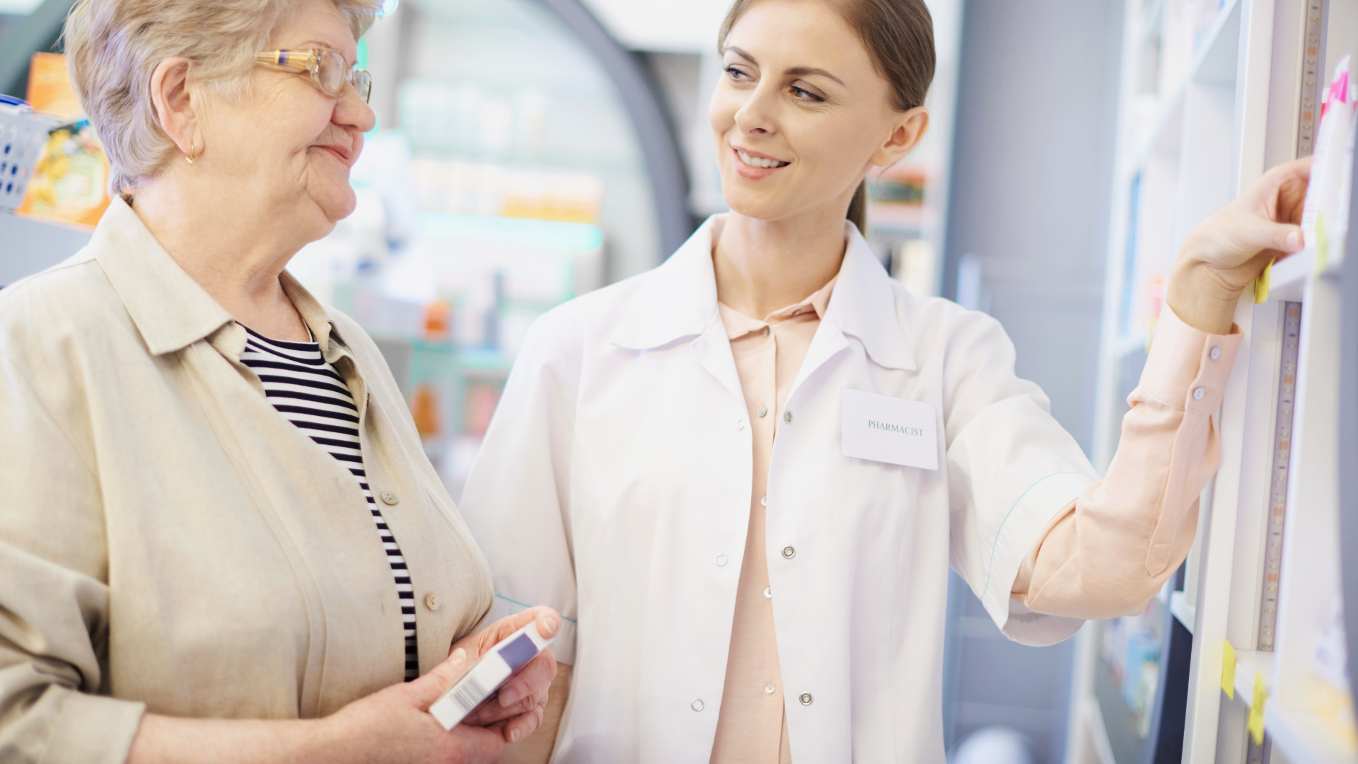 Zdjęcie przedstawia dwie kobiety w aptece. Starsza kobieta w okularach po lewej stronie trzyma w ręku pudełko z lekami. Po prawej znajduje się młoda, uśmiechnięta kobieta. Kobiety stoją obok regału w aptece.
