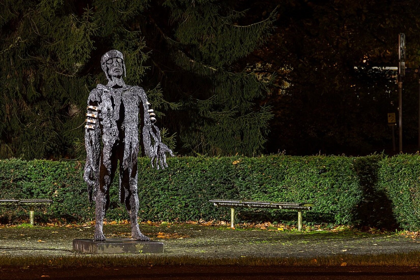 Zdjęcie przedstawia metalową rzeźbę. Przedstawia mężczyznę stojącego w lekkim rozkroku. Tułów rzeźby jest przecięty od klatki piersiowej w dół. Postać ma wyciągnięte do przodu ręce z dużymi dłońmi, głowę z założonym hełmem odchyla lekko w tył. W niedalekim odstępie otacza ją ciemnozielony żywopłot. Za mężczyzną stoją dwie ławki.