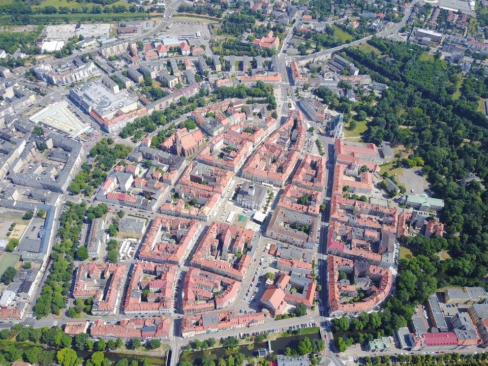 Zdjęcie przedstawia widok na miasto z lotu ptaka. Centrum miasta to podobne budynki z pomarańczowymi dachami. Stoją w zabudowie przypominającej kształtem prostokąty. Przedzielone są ulicami. W środkowej części starówki, pomiędzy budynkami, stoi biały obiekt z wieżą. Dookoła starówki rosną drzewa, a dalej są domy i bloki.      