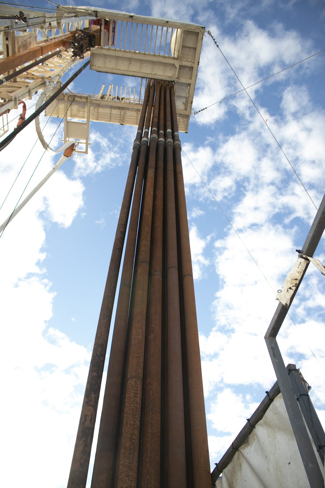 Na zdjęciu widoczne są od dołu rury płuczkowe, które składowane są pionowo na wiertni. Rury płuczkowe stanowią główną część przewodu wiertniczego. Służą one do przekazywania ruchu na narzędzie wiertnicze oraz do transportu płuczki wiertniczej na dno otworu wiertniczego. W lewym górnym rogu zdjęcia znajduje się biały maszt z przyległą do niego platformą. Maszt jest konstrukcją kratową o kształcie ostrosłupa. Platforma ma kształt dużej litery U/ Przez środek platformy poprowadzone są rury płuczkowe. Rury składowane są obok siebie w dwóch rzędach. Każda z rura jest rdzawego koloru. W środku każdej z rur znajduje się srebrne walcowate zgrubienie. W tle zdjęcia widać niebo.
Rury płuczkowe są podstawową częścią zestawu przewodu wiertniczego. Wykonane są jako stalowe rury bez szwu. Do każdego z końców rury przymocowany jest za pomocą zgrzewania tarciowego zwornik. Jest on częścią rury płuczkowej o zwiększonej średnicy zewnętrznej przez spęczenie. Na jednym końcu rury zgrzany jest zwornik zakończony gwintem narzędziowym wykonanym w postaci czopa, zaś na drugim końcu w postaci mufy. Rury płuczkowe ze względu na bardzo trudne warunki, w jakich pracują, wykonywane są ze specjalnych gatunków stali o dużej wytrzymałości. W kolumnie rur płuczkowych w czasie pracy występują naprężenia rozciągające (pochodzące od własnego ciężaru i ciężaru obciążników), naprężenia skręcające pochodzące od przenoszonego momentu obrotowego oraz naprężenia zginające. Oprócz tego wewnątrz rur płuczkowych przepływa płuczka, która wytwarza ciśnienie rozrywające na materiał rur oraz na połączenia zwornikowe.