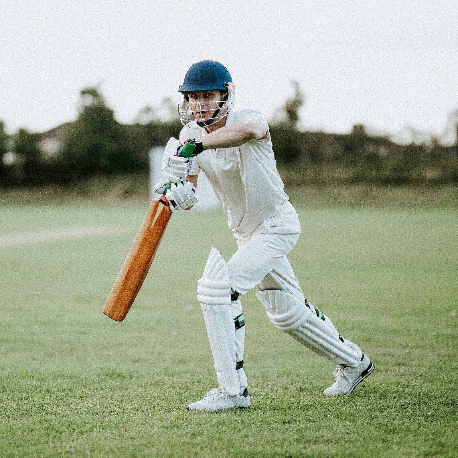 Zdjęcie przedstawia sportowca ubranego w biały strój sportowy i kask z siatką na twarz. Trzyma on w dłoni drewniany kij do krykieta.
