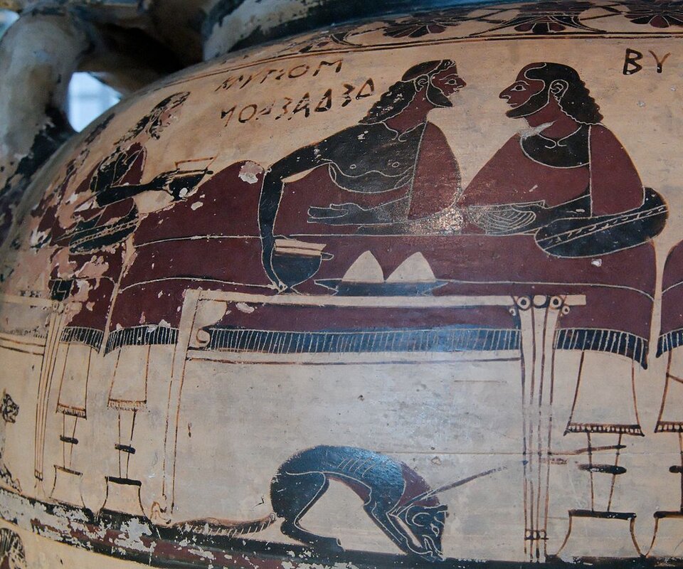 Na fotografii widoczny jest fragment wazy greckiej z namalowaną sceną biesiady. Dwóch brodatych mężczyzn półleżąc rozmawia podczas uczty. Z lewej strony widoczny jest mężczyzna, który przynosi nowe potrawy. Pod stołem skulony pies wyjada resztki. W górnej części wazy widoczne są zdobienia i napisy.