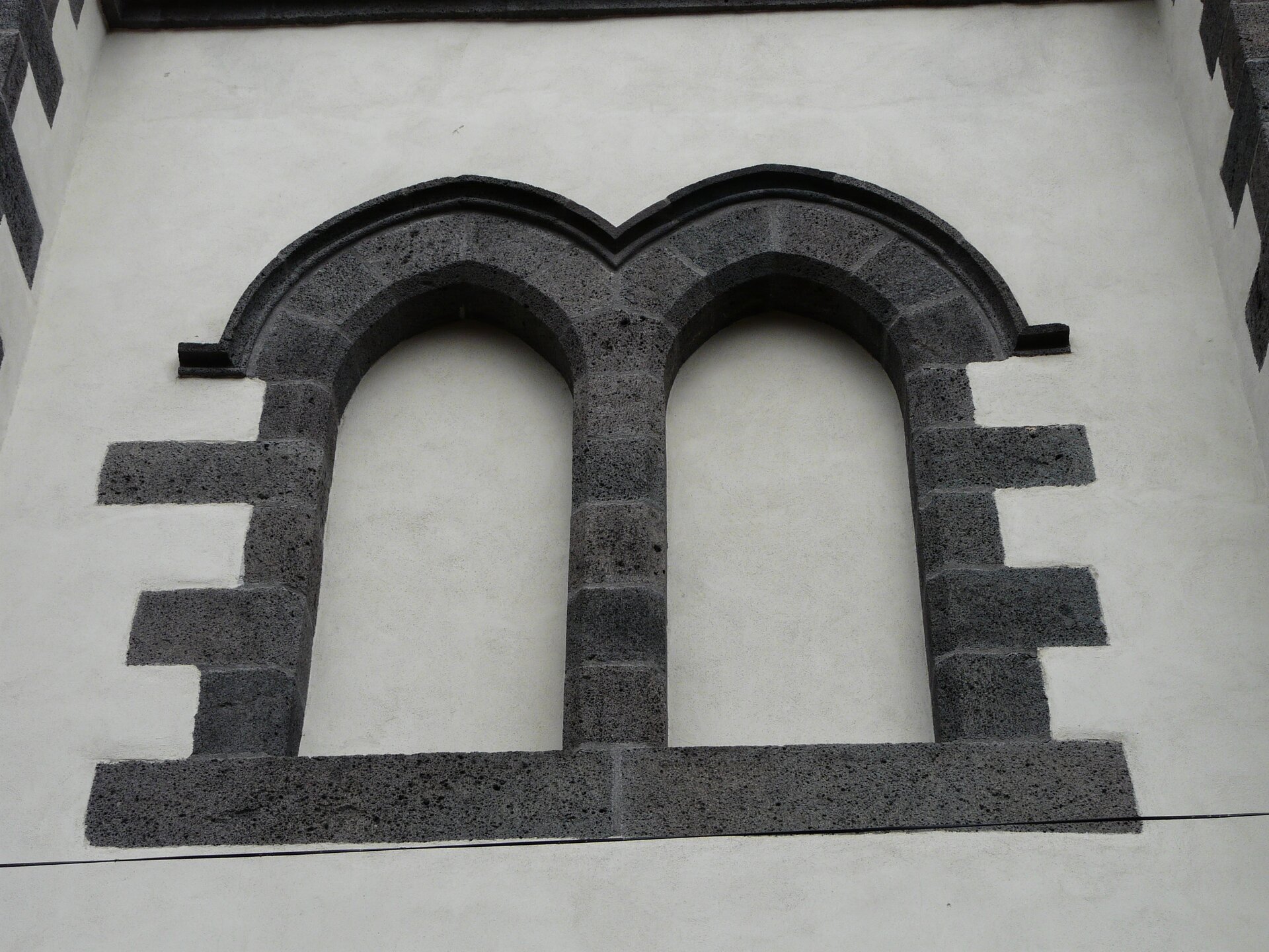 Fotografia przedstawia szarą elewację budynku, na której znajdują się dwa zaślepione otwory okienne, które zostały wykonane z ciemno szarej cegły i kontrastują z jasną gładką elewacją budynku. Okiennice mają kształt od góry zaokrąglonych prostokątów. 