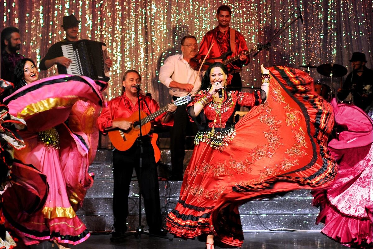 Zdjęcie przedstawia zespół cygański podczas koncertu. Z przodu kobiety podczas tańca, szerokie spódnice powiewają a ubrana w kolorowy, błyszczący strój solistka śpiewa do mikrofonu. Pomiędzy kobietami stoi mężczyzna, śpiewa i gra na gitarze.  Za nimi na podwyższeniu kilku mężczyzn  gra na instrumentach, skrzypcach, gitarze i perkusji. Z tyłu rozciąga się srebrzysta, pomarszczona kotara. 