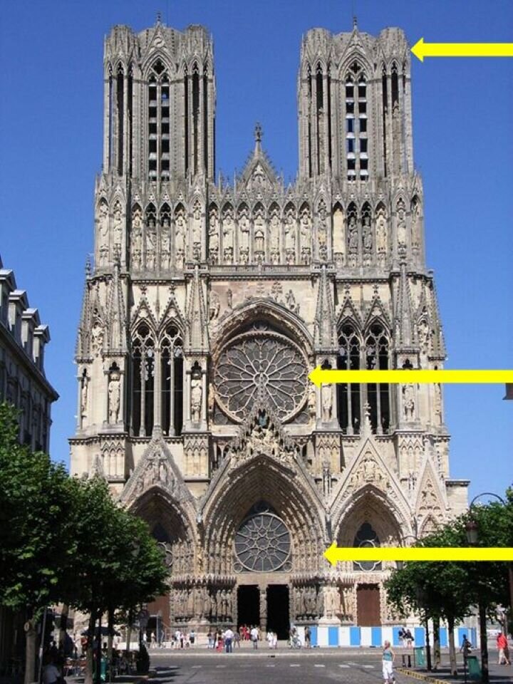 Ilustracja przedstawia budynek gotyckiej katedry w Reims we Francji. Budowla pochodzi z XIII wieku. Ukazuje widok od strony zachodniej z trzema portalami, rozetą nad wejściem głównym oraz bogatą dekoracją w postaci galerii królów. Katedra posiada dwie wysokie wieże. Fotografia ukazuje bryłę kościoła od strony wejścia, z drzewami rosnącymi po bokach. Tłem jest niebieskie niebo.