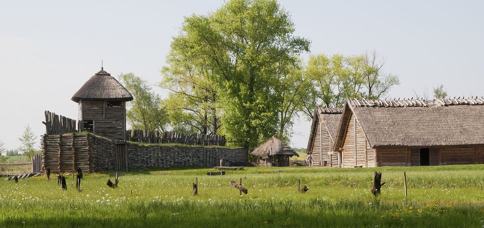 Na zdjęciu znajduje się fragment zrekonstruowanego grodu. Przedstawia ono drewniane umocnienie z wieżą wartowniczą oraz podłużne drewniane chaty za którymi rosną drzewa.