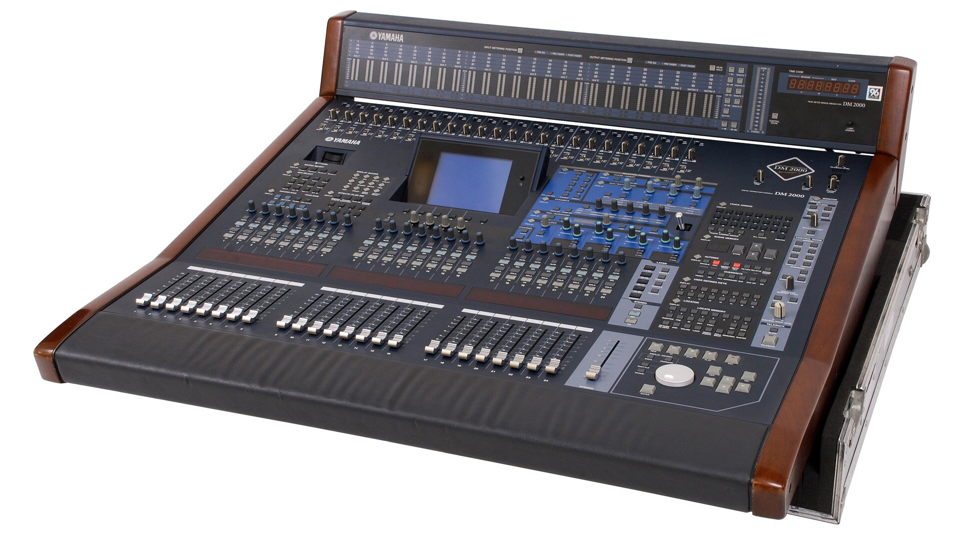 Na zdjęciu widoczny jest mikser cyfrowy. Ma kształt czarnego prostopadłościanu w ujęciu trzy czwarte, z odchylonym do góry tylnym, krótszym bokiem. Na urządzeniu widoczna jest duża ilość suwaków, pokrętem, lampek sygnalizacyjnych, przycisków. W górnej części widoczny niebieski prostokąt wpisany w czarny prostokąt – ekran, na prawo od którego niebieski prostokąt z dużą ilością przycisków i pokręteł.