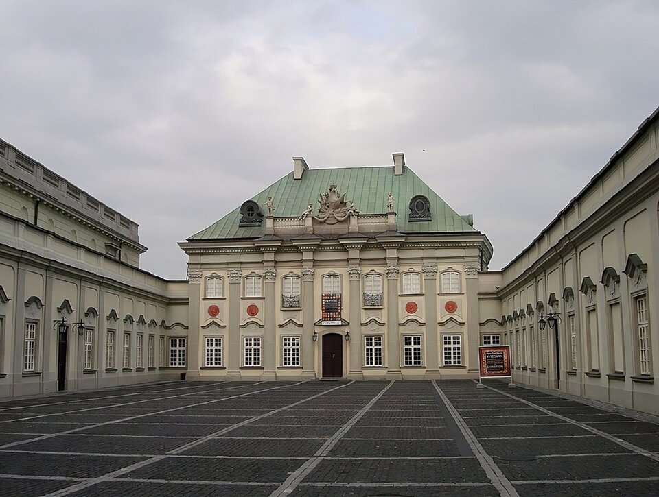 Zdjęcie przedstawia dwukondygnacyjny budynek od strony Zamku Królewskiego w Warszawie. Elewacja frontowa ozdobiona jest czterema kolumnami korynckimi i pilastrami. Pokryty miedzianą blachą dach korpusu pałacu jest czterospadowy z lukarnami. Na dachu, nad wejściem znajduje się rzeźba. Skrzydła boczne posiadają płaskie dachy i wysokie okna. Pomiędzy skrzydłami znajduje się brukowany kostką dziedziniec. 